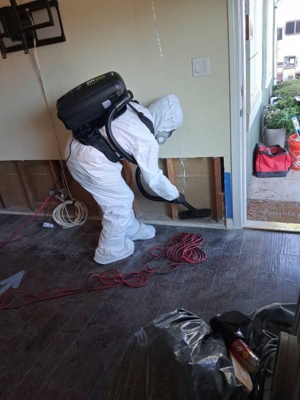 A technician with a backpack vacuum beside an opened wall over dark hardwood in Ventura County