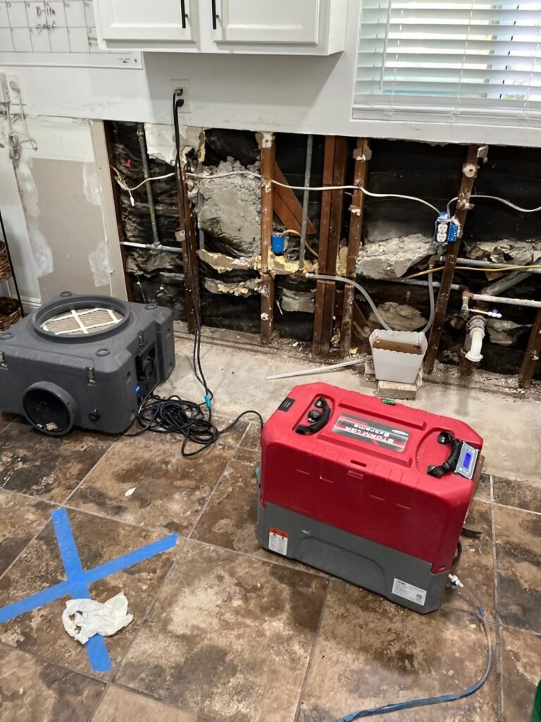 A kitchen wall opened with the plumbing exposed and two machines drying it