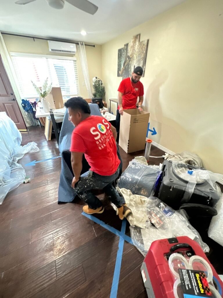 Two SOS technicians packing contents in a furnished living room with a dehumidifier running